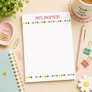 May include: A white rectangular notepad featuring the text Mrs. Morgan in pink at the top, bordered by horizontal rows of small multicolored dots in pink, yellow, green, and teal at both the top and bottom edges of the paper sheet.
