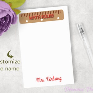 May include: White notepad with a brown ruler design at the top and the words "MATH RULES" in red. The notepad has the name "Mrs. Birdsong" in red script at the bottom. A clear pen rests on the side, with a purple flower in the background.