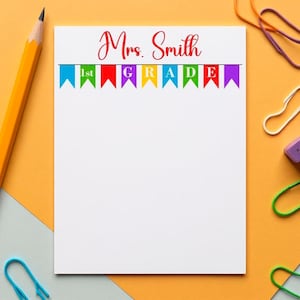May include: White notepad with the text "Mrs. Smith" in red script above a row of colorful pennants that spell out "1st GRADE". A yellow pencil and colorful paperclips are in the background.