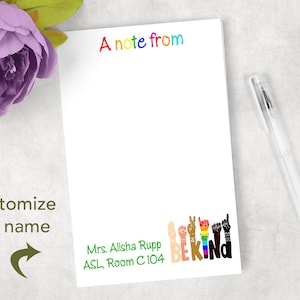 May include: White notepad with the text "A note from" in rainbow colors. The bottom of the notepad features the words "BE KIND" in colorful letters and hand gestures. A clear pen rests on the right side of the notepad.