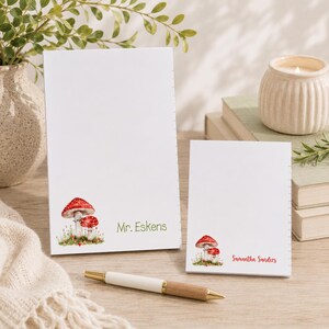May include: Two white notepads with a watercolor illustration of red mushrooms and green grass. One notepad has the name "Mr. Eskens" in green, and the other has "Samantha Sanders" in red. A pen rests in front of the notepads.