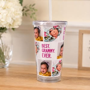 May include: A clear plastic tumbler with a straw featuring a collage of baby photographs and the pink text "BEST. GRAMMY. EVER." printed on the front. The cup has a transparent lid and a cylindrical shape, displaying multiple images of an infant.