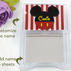 May include: A clear plastic dispenser with a Mickey Mouse design on the top. The design features red and white stripes, black ears, and the name "Chris" in yellow letters. The dispenser holds blank sheets of paper.