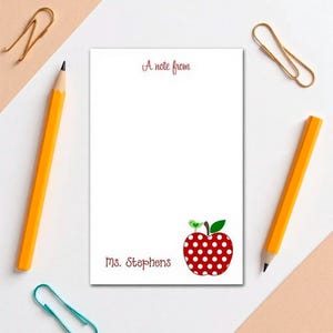 May include: White notepad with the text "A note from" and "Ms. Stephens" in burgundy script. A red apple with white polka dots and a green bird and leaf design is in the lower right corner. Two yellow pencils and gold paper clips are on the white surface.