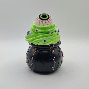 May include: A black plastic cauldron filled with bright green whipped topping. A large, realistic eyeball with a green iris sits on top, while smaller googly eyes and tiny orange, purple, and green sprinkles decorate the green topping.