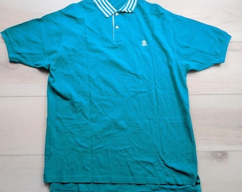 Puede incluir: Un polo de manga corta en color verde azulado con un cuello a rayas blancas y verde azuladas, una tapeta con tres botones y un pequeño emblema bordado en blanco en el pecho izquierdo. La prenda tiene un dobladillo recto y puños acanalados.