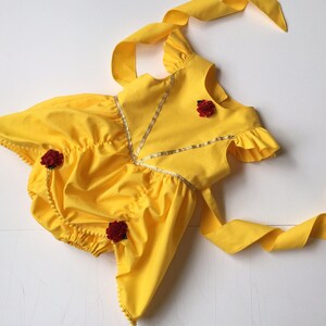May include: A bright yellow romper featuring ruffled sleeves, a V-shaped gold trim accent on the bodice, and three small dark red fabric roses attached to the front. The garment includes long yellow fabric ties at the shoulders and back.