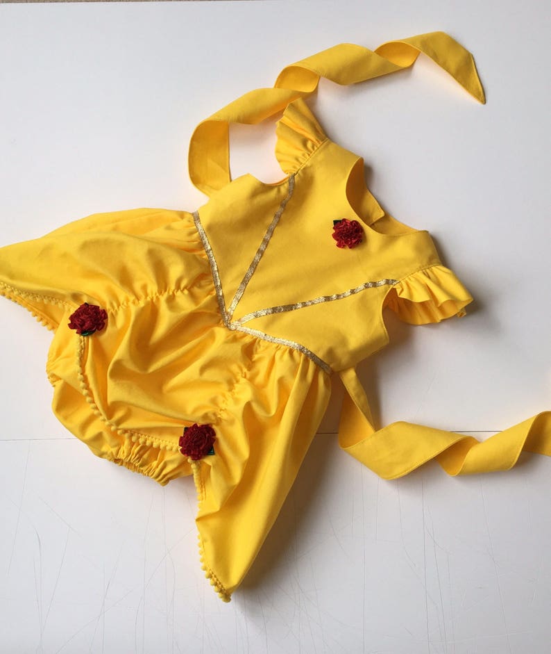 May include: A bright yellow romper featuring ruffled sleeves, a V-shaped gold trim accent on the bodice, and three small dark red fabric roses attached to the front. The garment includes long yellow fabric ties at the shoulders and back.