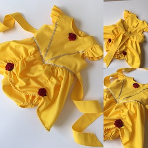 May include: A bright yellow romper featuring ruffled sleeves, a V-shaped gold trim across the bodice, and three small red fabric roses. The back includes a large tie-waist sash and three yellow buttons. The leg openings are gathered with elastic.