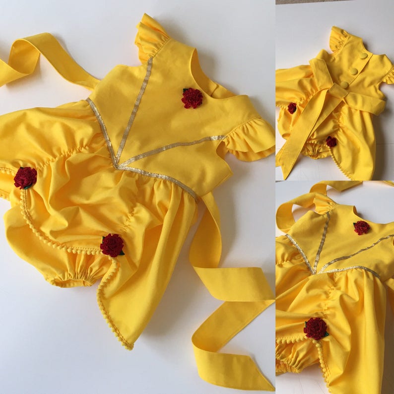 May include: A bright yellow romper featuring ruffled sleeves, a V-shaped gold trim across the bodice, and three small red fabric roses. The back includes a large tie-waist sash and three yellow buttons. The leg openings are gathered with elastic.