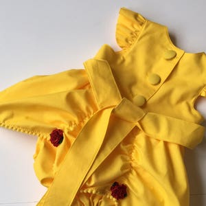 May include: A bright yellow romper featuring ruffled sleeves, a back button closure with three fabric-covered buttons, and a wide waist tie. The garment is accented with small red floral embellishments and delicate pom-pom trim along the leg openings.
