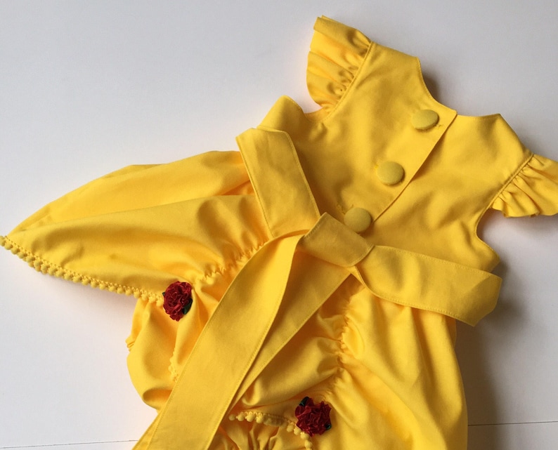 May include: A bright yellow romper featuring ruffled sleeves, a back button closure with three fabric-covered buttons, and a wide waist tie. The garment is accented with small red floral embellishments and delicate pom-pom trim along the leg openings.