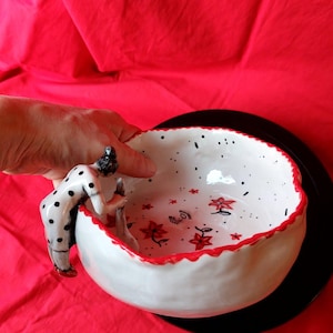 Può includere: Una ciotola in ceramica bianca con bordo smerlato rosso, decorata con pois neri e illustrazioni di fiori rossi. Una figura scolpita con un vestito a pois bianchi e neri è fissata sul lato, intenta a mescolare l'interno della ciotola.