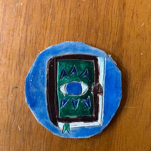 May include: A circular, hand-painted leather patch featuring a green book with a central blue eye illustration. The book is framed by a dark brown border and set against a vibrant blue background, with small blue triangular accents surrounding the eye.