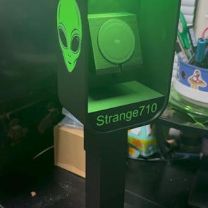 May include: A dark green, rectangular phone booth stand with a black base and post. The top section features the word PHONE in white text, a green alien head graphic on the side, and the text Strange710 on the front, with a wireless charger inside.