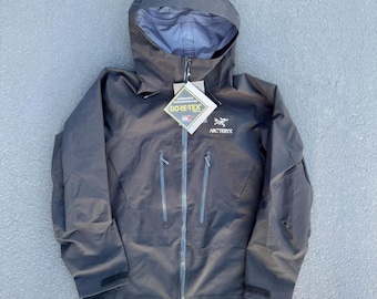 Può includere: Una giacca nera con cappuccio Arc'teryx dotata di cerniera frontale a tutta lunghezza, due tasche verticali sul petto con cerniere e polsini regolabili. Un'etichetta del marchio Gore-Tex è attaccata sul davanti e il logo Arc'teryx è stampato sul petto.