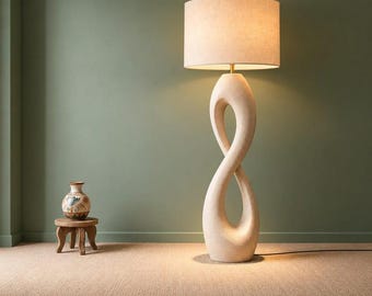 Może przedstawiać: Lampa podłogowa z beżową, teksturowaną podstawą w kształcie nieskończoności i cylindrycznym abażurem z tkaniny. Obok znajduje się mały, okrągły drewniany stołek z ozdobnym ceramicznym wazonem w brązowe i czarne wzory.