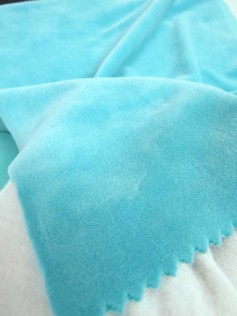 May include: A close-up shot of a soft, light blue fabric with a velvety texture. The fabric is folded, displaying different shades of blue. The edge of the fabric has a decorative, scalloped trim. The fabric is likely used for clothing or home decor.