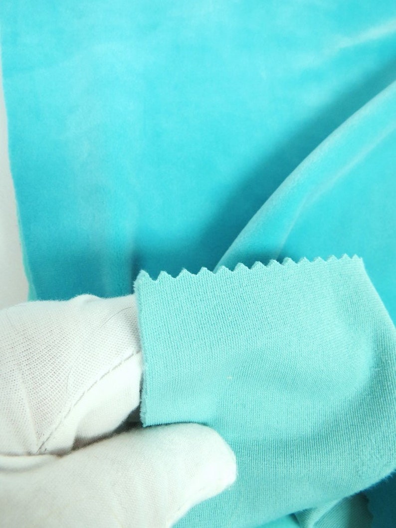 May include: Close-up of a vibrant turquoise fabric. The fabric has a soft, velvety texture. A section of the fabric is held up, revealing a serged edge. The fabric is a solid colour and appears to be a knit or jersey material.