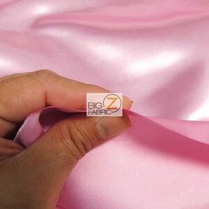 May include: Close-up of a hand holding a section of pink satin fabric. The fabric has a smooth, shiny texture and is folded in soft waves. The logo "BIG Z FABRIC" is visible in the upper right corner.