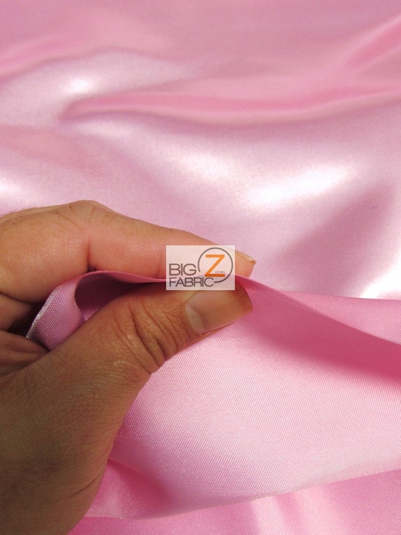 May include: Close-up of a hand holding a section of pink satin fabric. The fabric has a smooth, shiny texture and is folded in soft waves. The logo "BIG Z FABRIC" is visible in the upper right corner.