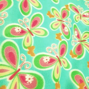 Op de afbeelding: Een zachte stof met een herhalend patroon van vlinders op een heldere turquoise achtergrond. De vlinders hebben roze centra, gele en groene vleugels en witte bloemaccenten. Kleine oranje vlindersilhouetten zijn verspreid over het geheel.
