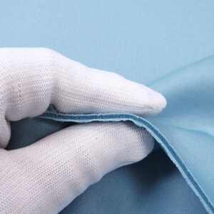 May include: A close-up of a light blue fabric with a white glove holding it up. The fabric is smooth and has a slight sheen.