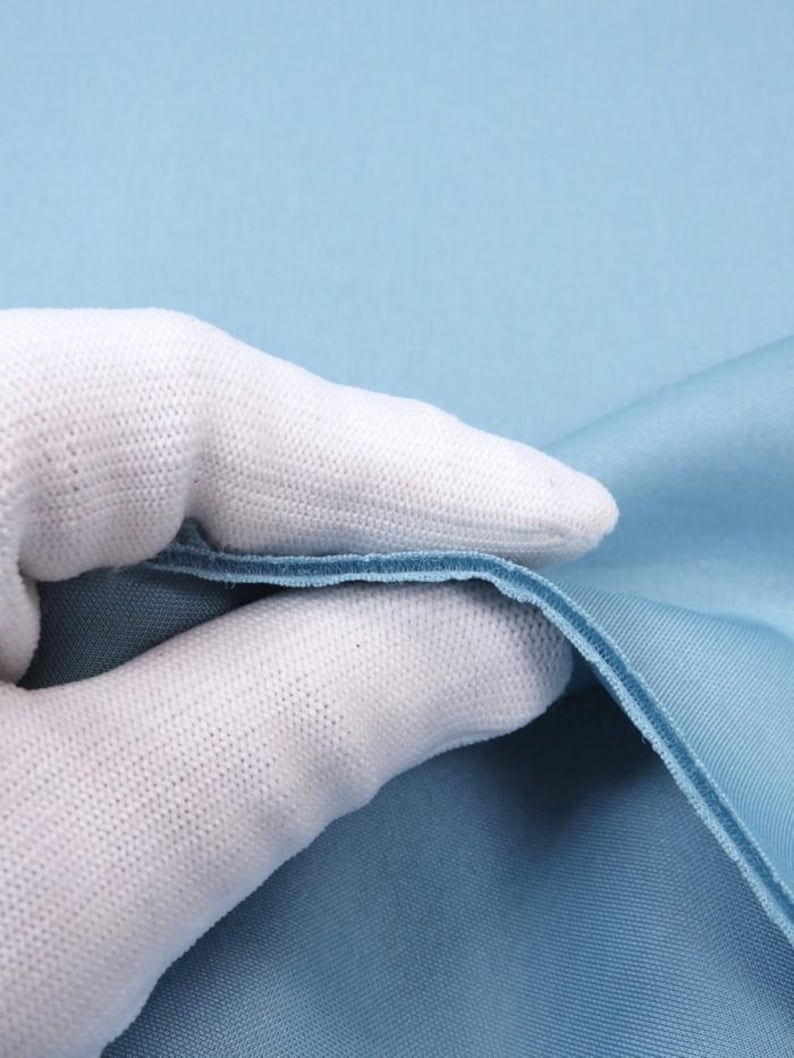 May include: A close-up of a light blue fabric with a white glove holding it up. The fabric is smooth and has a slight sheen.