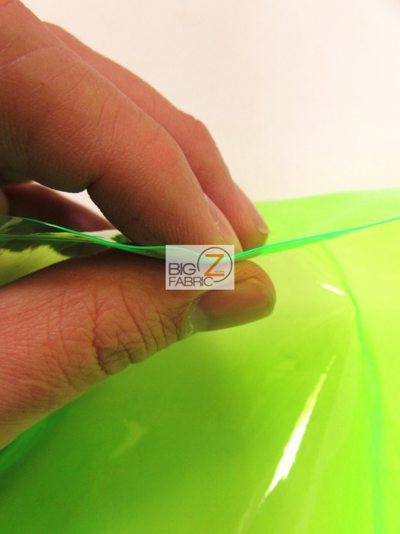 Puede incluir: Primer plano de una mano tocando una l&aacute;mina de tela verde ne&oacute;n brillante. La tela es transl&uacute;cida y refleja la luz. El logotipo "BIG Z FABRIC" es visible en la esquina.
