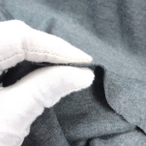 May include: Close-up of a dark gray fabric with a soft, textured surface. The fabric is folded and held by a white gloved hand.