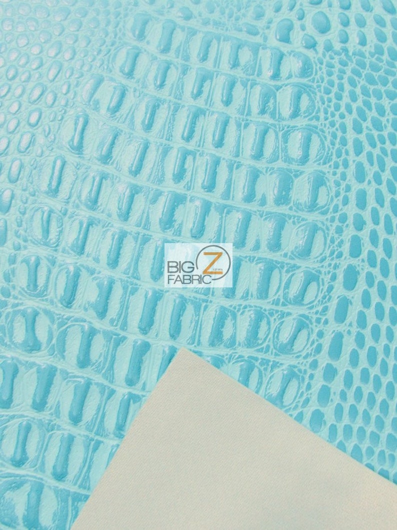 Op de afbeelding: Close-up van een lichtblauwe kunstleer met krokodil-embossing. De stof heeft een textuur met schubben. De stof wordt getoond naast een stuk witte stof.