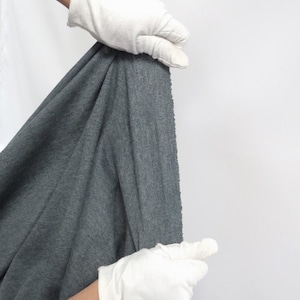 May include: A close-up of a dark gray fabric being held by gloved hands. The fabric has a textured surface and a slightly rippled appearance.