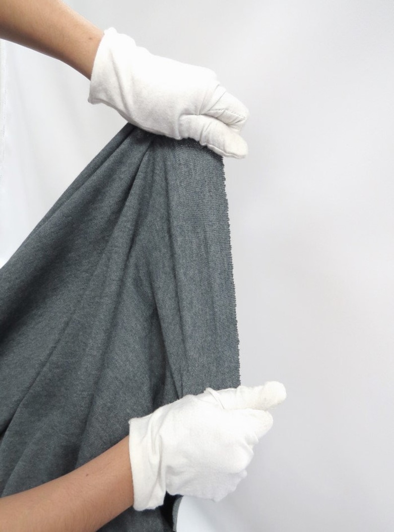 May include: A close-up of a dark gray fabric being held by gloved hands. The fabric has a textured surface and a slightly rippled appearance.