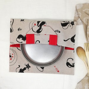 Puede incluir: Una bolsa de tela beige con diseños de gatos negros y asas rojas. Una bandeja para hornear de metal plateado está dentro de la bolsa.