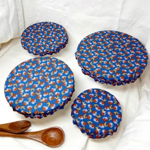 Könnte beinhalten: Vier blaue und weiße Stoffabdeckungen mit einem floralen Muster in Orange- und Weißtönen. Die Abdeckungen sind in verschiedenen Größen und auf einer weißen Oberfläche angeordnet.