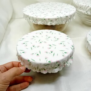 Op de afbeelding: Drie witte stoffen kom deksels met een roze bloemenpatroon. De deksels zijn gemaakt van een zachte, ademende stof en zijn perfect om voedsel vers te houden.