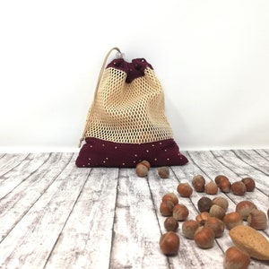 May include: A beige mesh drawstring bag with a burgundy base and trim, dotted with small white circles. The bag is filled and sits on a wooden surface scattered with hazelnuts and an almond.