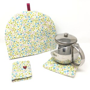 Può includere: Un set di tre accessori per teiera con un motivo floreale. La teiera è in tessuto bianco con un motivo floreale blu e verde. La teiera ha un nastro rosa. La teiera è su una superficie bianca. C'è una teiera abbinata e un porta bustine di tè.