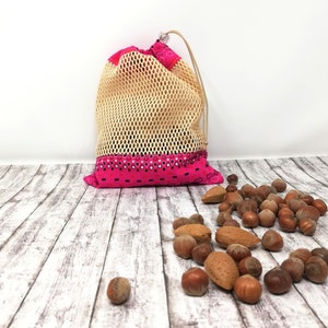 May include: A pink and beige mesh drawstring bag with a white background and a pink and black patterned border. The bag is sitting on a whitewashed wood surface with a pile of hazelnuts and almonds scattered around it.