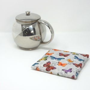 Puede incluir: Una tetera plateada con cuerpo de vidrio y un calentador de té con estampado de mariposas.