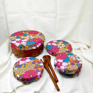 Peut inclure: Quatre couvercles de bol en silicone réutilisables avec un motif floral dans des tons de rose, jaune, bleu et orange. Les couvercles sont sur des bols en bois. Deux cuillères en bois sont également incluses.