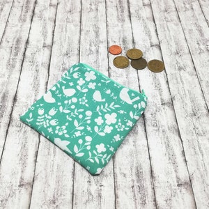 May include: A small teal zippered pouch with a white floral and bird pattern. The pouch is lying on a whitewashed wood surface.