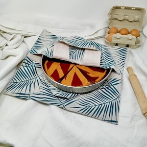 Puede incluir: Una tarta con relleno rojo y corteza decorativa está en una sartén redonda de metal, cubierta por una tela con estampado de hojas de palmera verde azulado y blanco. También hay una caja de huevos y un rodillo de madera.