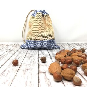 May include: A yellow mesh drawstring bag with a blue and white patterned bottom. The bag is sitting on a whitewashed wood surface with a pile of almonds and hazelnuts scattered around it.