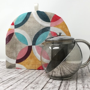 May include: A silver teapot with a glass body and a stainless steel lid and handle. The teapot is sitting on a wooden surface next to a colorful tea cozy with a geometric pattern.