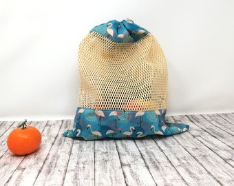 May include: A beige mesh drawstring bag with a blue and white flamingo print lining. The bag is sitting on a wooden surface with a small orange.