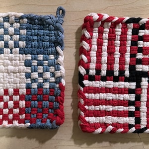 May include: Two woven potholders in red, white, and blue. One features a design reminiscent of the American flag, while the other has a similar pattern with black accents. Both have a loop for hanging.