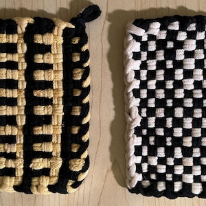 May include: Two woven potholders. One is black and tan with vertical stripes. The other is black and white checkered. Both have a loop for hanging. Kitchen and home decor.