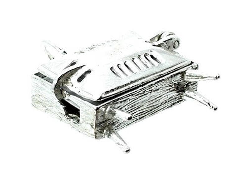 May include: A small, silver-colored miniature barbecue grill with a hinged lid and four legs. The grill has a textured surface and decorative details, including a handle and vents on the lid. The overall design suggests a novelty or decorative item.
