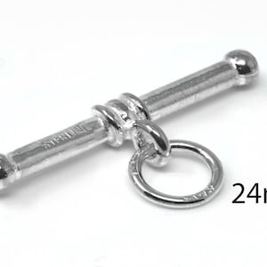 Op de afbeelding: Een zilverkleurige toggle sluiting, 24 mm lang, met een staaf- en ringontwerp. De staaf heeft afgeronde uiteinden en een gestructureerd midden. De ring is met een kleine lus aan de staaf bevestigd. Het woord "STERLING" is zichtbaar op de staaf.